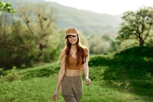 un hermosa mujer en ropa de deporte y un gorra camina en contra un verde natural paisaje y sonrisas en el luz de sol foto