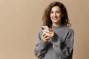 Happy curly beautiful lady in gray casual sweater chatting with boyfriend looks at camera posing isolated on beige pastel background. Social media, network, distance communication concept. Copy space photo