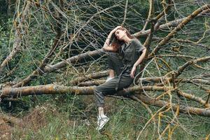 A woman in a green jumpsuit touches her head and sits on a broken tree photo