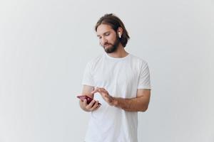 A man with a beard in white T-shirt and jeans looks at his phone flipping through an online social media feed with headphones in his ears listening to a voice message on a white background and smiling photo