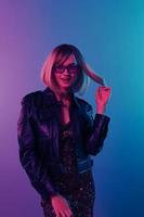 Flirting sexy smiling beautiful blonde woman in leather jacket sparkly dress trendy sunglasses posing isolated in blue pink color light studio background. Neon party Cyberpunk concept. Copy space photo