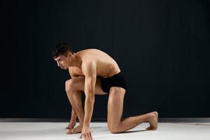 handsome man muscularly muscled in black panties stands on his knee dark background photo