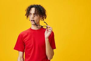 Incredulous myopic young student man in red t-shirt hold funny eyewear on teeth posing isolated on yellow studio background. The best offer with free place for advertising. Education College concept photo