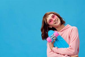 Cute smiling pretty redhead lady in pink hoodie sunglasses with penny board look at camera posing isolated on blue studio background. Copy space Banner Offer. Fashion Cinema. Holiday activity photo