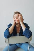 A young woman sitting in a chair at home smiling with teeth with a short haircut in jeans and a denim shirt on a white background. Girl natural poses with no filters photo