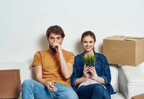 man and woman sitting on a white sofa in a room with boxes of things tools moving photo