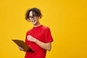 Joyful happy myopic young student man in red t-shirt funny eyewear holds tablet folder with study notes posing isolated on over yellow studio background. Free place for ad. Education College concept photo