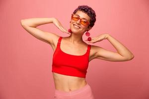 Young athletic woman with a short haircut and purple hair in a red top and pink yoga leggings in sunglasses with an athletic figure smiles and dances on a pink background photo