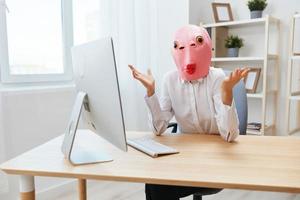 Pensive crazy businesswoman worker freelancer in pink fish mask work on computer online pondering or making decision thinking of problem solution spread hands in light modern office. Copy space photo