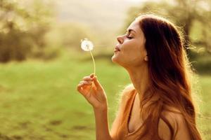 retrato de un joven mujer en perfil con un diente de león flor en su mano soplo en eso y sonriente en contra el verde verano césped en el ajuste Dom en naturaleza foto