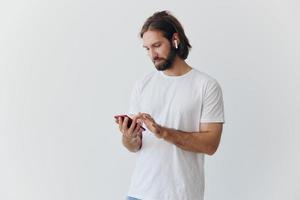 A man with a beard and long hair in a white T-shirt and blue jeans looks at his phone flipping through an online social media feed with headphones in his ears against a white background photo