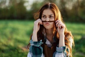 gracioso mujer retrato de cerca riendo y meneando para el cámara en frente de un verde parque en verano, el puesta de sol brillante en su rojo pelo foto