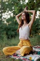 A young hippie woman meditates in nature in the park, sitting in a lotus position on her colorful plaid and enjoying harmony with the world in eco-clothing photo