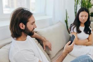 A man looks at his phone screen and smiles during an argument with his girlfriend. The angry and hurt woman looks in his direction and is sad. Family discord at home, phone addiction photo