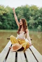 A hippie woman sits on a bridge by a lake with her hands up in the air while traveling in nature and smiling in eco-clothing. Relaxed lifestyle photo