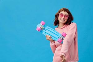 Happy cheerful pretty redhead lady in pink hoodie sunglasses with penny board posing isolated on blue studio background. Copy space Banner Offer. Fashion Cinema. Holiday activity concept photo