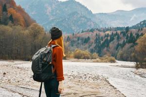 un viajero con un mochila en un suéter y pantalones son descansando en el montañas en naturaleza cerca el río en otoño foto