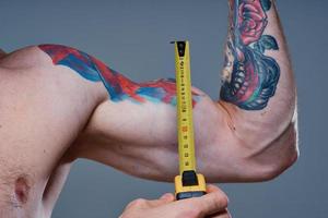 guy measures arm muscles with centimeters on a gray background and a multicolored tattoo bodybuilder fitness photo