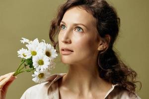 Close up portrait of adorable curly female in linen casual shirt hold chamomiles flowers near face looks up posing isolated on over olive green pastel background. Nature is beauty concept. Copy space photo