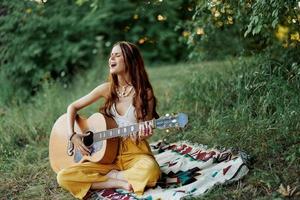 joven hembra hippie artista obras de teatro guitarra y canta canciones en Respetuoso del medio ambiente ropa sentado en el suelo fuera de en naturaleza en el otoño mirando fuera a el puesta de sol foto