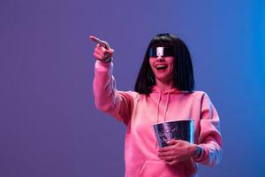 Laughing awesome brunet woman in pink hoodie trendy specular sunglasses with popcorn point finger aside posing isolated in blue violet color light background. Neon party Cinema concept. Copy space photo