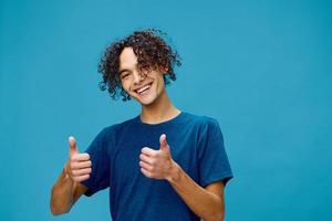 Cheerful happy curly tanned Caucasian guy in basic t-shirt show both thumbs up gesture Like posing isolated on over blue background. Lifestyle and Emotions concept. Good offer with copy space for ad photo