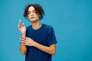 Funny cute curly tanned Caucasian guy in basic t-shirt folds hands looks at camera posing isolated on over blue background. Lifestyle and Emotions concept. Good offer with copy space for ad photo