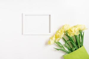 A green paper bag with spring flowers of yellow tulips and a white empty text frame on a white background. Flat layout, top view, copy space, mockup photo