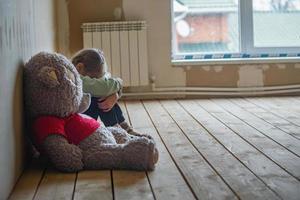 a nine-year-old girl is upset, she is sitting in a room without repairs on the floor, hugging her knees and crying, next to a large soft toy bear, copy space photo