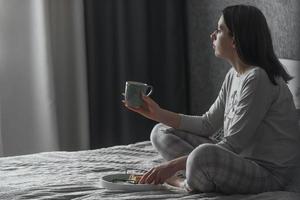 un hermosa joven niña en pijama se sienta solo en el cama en el Mañana en su dormitorio, en su manos es un jarra de té, ella mira fuera el ventana foto