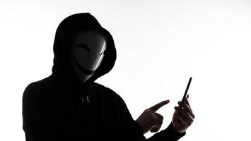 hacker anónimo y mascarilla con smartphone en la mano. hombre con camisa de capucha negra sosteniendo y usando un teléfono móvil con fondo blanco. representan el concepto de piratería informática o robo de datos personales foto