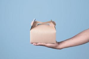 Male hand holding brown ecological package box made of natural cardboard on light blue background. Packaging, shopping, Food delivery concept photo