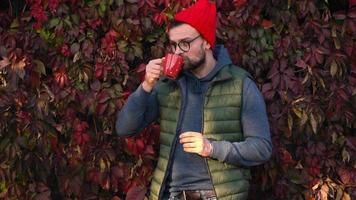 Mann im ein rot Deckel mit ein Tasse im seine Hände Trinken Tee oder Kaffee draußen. Kaffee Unterbrechung. video