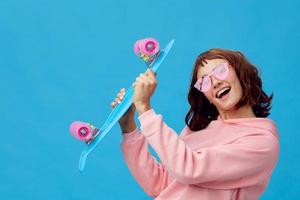 Happy cheerful pretty redhead lady in pink hoodie sunglasses with penny board posing isolated on blue studio background. Copy space Banner Offer. Fashion Cinema. Holiday activity concept photo