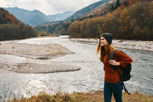 un mujer en un rojo chaqueta y un mochila en su espalda son caminando en el bancos de el río en el montañas foto