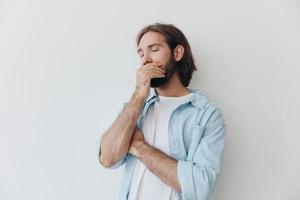 milenario hombre con un barba soportes en contra un blanco pared y cubre su cara con su mano desde tristeza, estrés foto