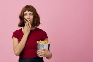 Excited shocked cute redhead lady in red t-shirt with popcorn ready for movie evening posing isolated on over pink studio background. Copy space Banner. Fashion Cinema concept. Entertainment offer photo