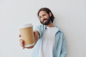 Lanza libre milenario hombre con un barba Bebiendo café desde un reciclado taza en elegante hipster ropa blanco camiseta azul pantalones y camisa en un blanco antecedentes foto