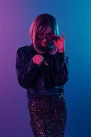 Confused sexy beautiful blonde woman in leather jacket sparkly dress trendy sunglasses point finger at camera posing isolated in blue pink color light studio background. Neon party Cyberpunk concept photo