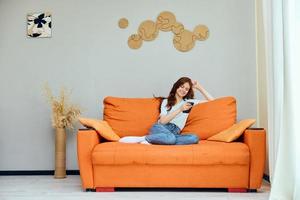 alegre mujer chateando en el naranja sofá con un teléfono inteligente apartamentos foto
