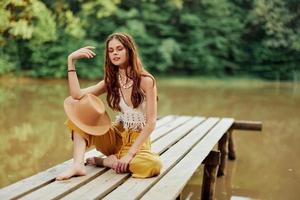 un joven hippie mujer se sienta en un lago puente vistiendo elegante eco ropa y sonriente foto