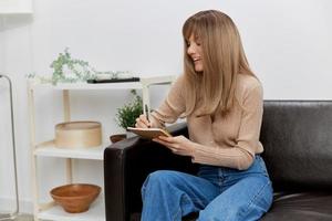 Focused cheerful blonde student lady with copybook enjoy cool task sitting in sofa couch at modern living room interior. Female client have shrink appointment in psychological help office. Copy space photo