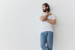 A man with a beard and long hair in a white T-shirt and blue jeans stands against a white wall, leaning against it and listening to music with wireless white headphones, staring thoughtfully photo