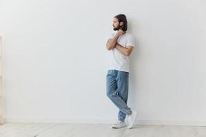 A man with a beard and long hair in a white T-shirt and blue jeans stands against a white wall leaning full-length on it, relaxed style photo