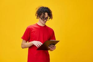 Confused myopic young student man in red t-shirt funny eyewear holds tablet folder with study notes posing isolated on over yellow studio background. Free place for ad. Education College concept photo