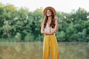 un joven mujer en un hippie Mira viajes en naturaleza por el lago vistiendo un sombrero y amarillo pantalones en el otoño foto