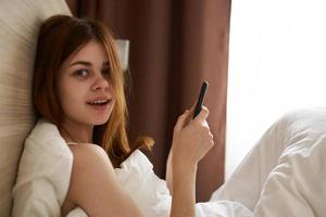 woman with mobile phone lies in bed and window blanket curtains photo