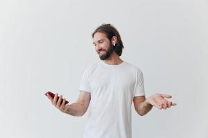 un hombre con un hipster barba en un blanco camiseta con un teléfono y inalámbrico auriculares sonriente escuchando a música y un audio libro en línea en contra un blanco pared foto