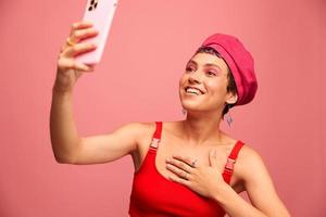 un joven mujer blogger con de colores rosado pelo y un corto Corte de pelo toma un imagen de sí misma en el teléfono y retransmisiones un sonrisa en elegante ropa y un sombrero en un rosado antecedentes monocromo estilo foto