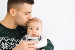 un joven padre sostiene su pequeño hijo en su brazos y Besos él en un blanco antecedentes foto